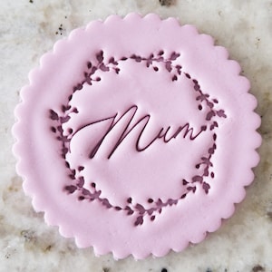 May include: A light pink round cookie with a scalloped edge, decorated with a floral wreath and the word "Mum" in cursive.