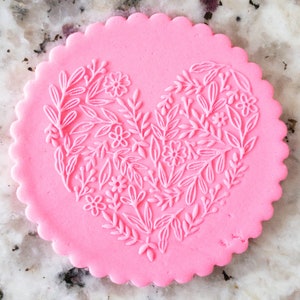 May include: A pink heart-shaped cookie with a white floral design embossed on the surface.