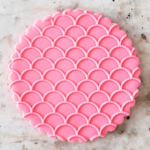 Peut inclure: Emporte-pièce rond en fondant rose avec un motif d'écailles de sirène. L'emporte-pièce a un bord festonné.