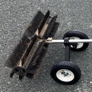May include: A wheeled push broom featuring a cylindrical rotating brush with dense black bristles, a silver metal handle, and two black pneumatic tires with white rims. The assembly includes a black support frame connecting the brush to the wheels.