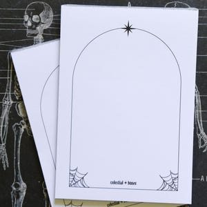 以下が含まれることがあります： 黒いアーチ型の縁、星、クモの巣の角のデザインが施された2つの白いメモ帳。「celestial + bones」の文字が下部にあります。背景には、骨格図と「SKELETAL SYSTEM」の文字があります。