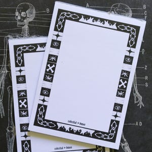 May include: Two white notepads with black decorative borders featuring celestial and bone-themed designs. The notepads have the text "celestial + bones" at the bottom. The background is a chalkboard-style illustration of the skeletal system.