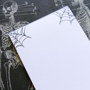 May include: A white notepad with black spiderweb designs in the corners, set against a vintage anatomical chart background. The notepad is blank, offering space for notes or sketches. The background features a skeletal illustration.