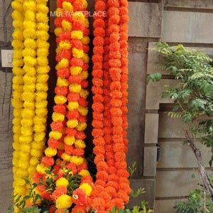Könnte beinhalten: Drei lange dekorative Girlanden aus künstlichen Ringelblumen, die vertikal hängen. Eine Girlande ist einfarbig gelb, eine ist eine Mischung aus Gelb und Orange und eine ist einfarbig orange. Der Text MULTIARTPLACE ist auf den Girlanden zu sehen.