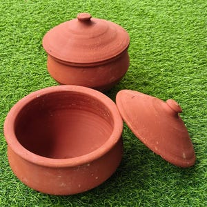 May include: Two terracotta clay pots with matching lids. One pot is shown with its lid placed securely on top, while the other pot is open with its lid resting beside it. The pots have a rounded shape, a wide opening, and a smooth, reddish-brown finish.