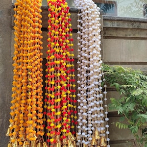 May include: Three vertical decorative garlands featuring strings of small pom-poms in solid yellow, a mix of red and yellow, and solid white. Each strand is interspersed with small gold-colored beads and finished with a gold-toned metal bell at the base.