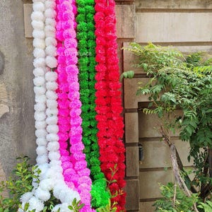 May include: Four long, vertical garlands of artificial flowers in white, bright pink, vibrant green, and red. Each garland consists of numerous densely packed, rounded, ruffled fabric blossoms strung together. The text MULTIARTPLACE is visible.