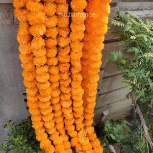 May include: A long decorative garland composed of multiple strands of vibrant orange marigold flowers. The individual blossoms are densely packed and rounded, creating a textured, cascading effect. The text multiartplace is visible.