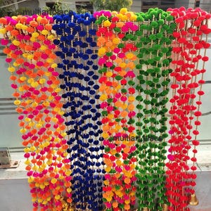 May include: A set of five vertical hanging garlands featuring rows of colourful fabric pom-poms in shades of pink, yellow, orange, blue, green, and red. Each strand is accented with small gold-toned beads and finished with a gold-toned metal bell.