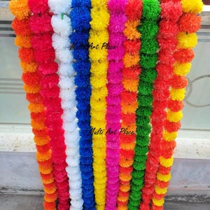 May include: A collection of vertical garlands made of artificial marigold flowers in vibrant colors including orange, red, white, blue, yellow, pink, and green. The text Multi Art Place is visible on the strands of blue and yellow flowers.