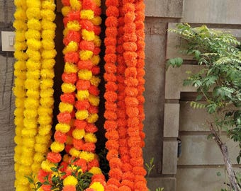May include: Three long decorative garlands made of artificial marigold flowers hanging vertically. One garland is solid yellow, one is a mix of yellow and orange, and one is solid orange. The text MULTIARTPLACE is visible on the garlands.