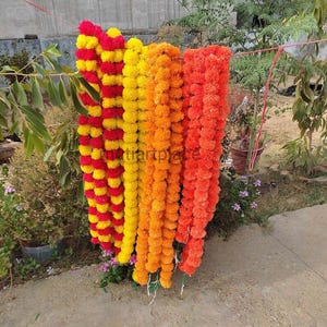 May include: A collection of long, hanging floral garlands made of artificial marigold flowers in vibrant shades of red, yellow, and orange. The garlands are suspended from a thin red string, displaying dense clusters of layered, ruffled petals.