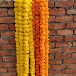 May include: A decorative garland consisting of multiple strands of artificial marigold flowers in vibrant yellow and orange hues. The flowers are densely packed, creating a full, textured appearance. The text "metiartplace" is visible in the center.