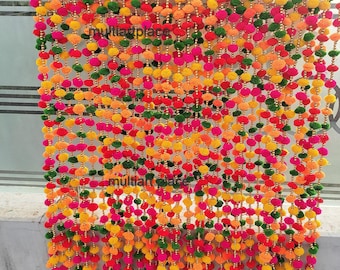 Puede incluir: Una cortina decorativa colgante compuesta por múltiples hilos verticales con pequeños pompones coloridos alternos en tonos rosa, amarillo, naranja y verde, intercalados con cuentas doradas y terminados con pequeñas campanas doradas.