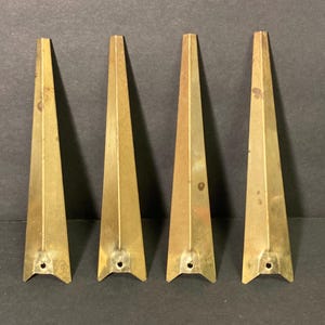 May include: Four brass-colored, triangular furniture feet. Each foot has a pointed top and a flared base with a small hole. The feet are arranged in a row against a dark background.