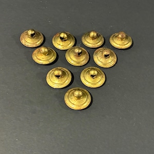 May include: Ten antique brass furniture knobs with a rounded top and a central hole. The knobs have a tarnished, aged appearance, suggesting they are vintage. They are arranged on a dark surface, showcasing their golden color and intricate design.