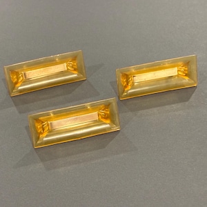 May include: Three rectangular, gold-colored decorative objects with a lighter gold center section. The objects are arranged on a gray surface. The objects have a slightly translucent appearance.