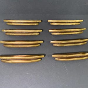 May include: Eight antique brass drawer pulls. Each pull has a curved, layered design with a warm, golden finish. The pulls are arranged in two rows of four against a dark background, suggesting a vintage or retro aesthetic.