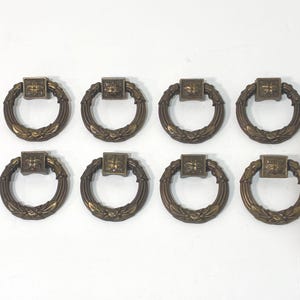 May include: Eight antique bronze drawer pulls. Each pull features a square center piece with a decorative design, connected to a circular handle with a leaf pattern. The pulls are arranged in two rows of four against a white background.