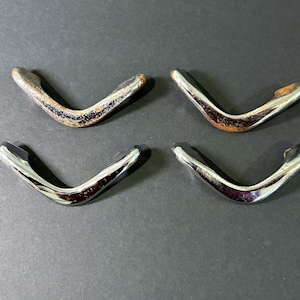 May include: Four V-shaped metal drawer pulls. Two are chrome, and two have a distressed, aged appearance with brown and black tones. The pulls are arranged on a dark gray surface.