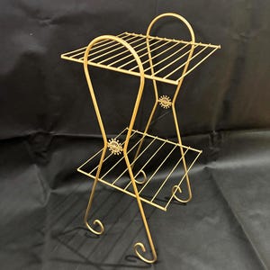 May include: A gold-colored, two-tiered metal plant stand. The stand features a rectangular top and bottom shelf with a slatted design. Decorative floral accents are present on the frame. The legs have a curved, scroll-like design.
