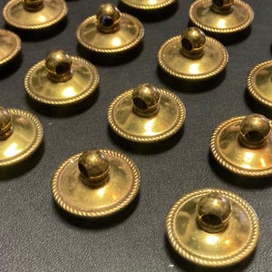 May include: A collection of antique brass buttons with a domed design and a small, round knob on top. The buttons have a decorative rope-like border and are arranged on a dark surface. These buttons are likely for clothing or other craft projects.