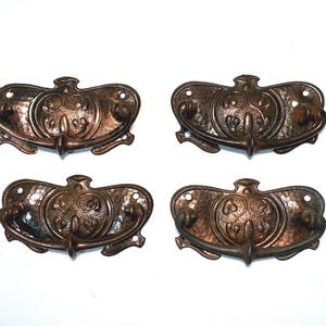 May include: Four antique bronze drawer pulls. Each pull has a hammered texture, a central decorative medallion, and a curved handle. The pulls are symmetrical and feature ornate detailing, perfect for vintage furniture restoration.