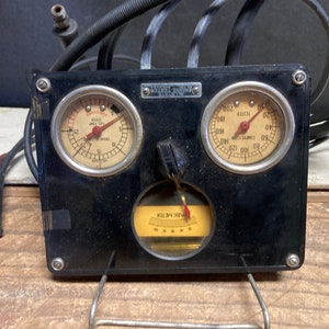 May include: Vintage black engine tester with two gauges and a spark meter. The gauges read "Fuel Pump Tester" and "Compression Tester". The spark meter is labeled "Spark Meter". The device has wires and a metal stand.