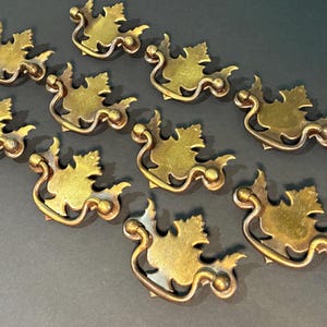May include: A collection of ten antique brass drawer pulls. Each pull features a leaf design with a curved handle. The brass has a warm, aged patina, suggesting vintage craftsmanship. Ideal for furniture restoration projects.