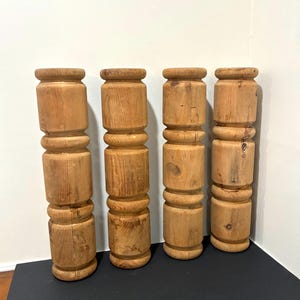May include: Four unfinished wooden decorative columns. Each column is cylindrical with a series of rounded sections. The wood grain is visible, and the columns are a light brown color. They are arranged vertically against a white wall.