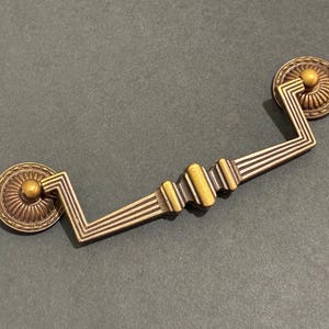 May include: A brass-toned metal drawer pull featuring a swinging handle with a stepped, geometric design. The handle is attached to two circular, fluted backplates with a central knob detail, showcasing a weathered, antique-style finish.