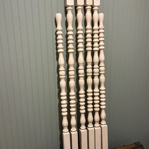 May include: A collection of seven antique, off-white wooden balusters. Each baluster features a turned design with a series of rounded and elongated sections. The balusters are of varying lengths and are arranged against a green-toned wall and a wooden trunk.