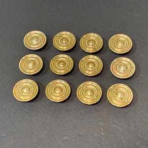 May include: A set of twelve circular gold-toned buttons arranged in a grid of three rows and four columns. Each button features a raised central dome surrounded by three concentric circular ridges, creating a tiered, metallic, reflective surface.