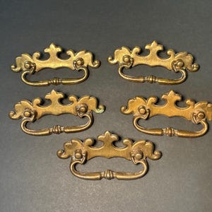 May include: Five antique brass drawer pulls with ornate, scroll-like designs. Each pull features a curved handle and decorative flourishes, perfect for vintage furniture restoration. The pulls are arranged on a dark gray surface.