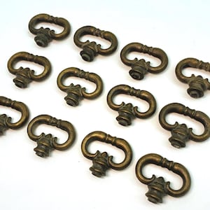 May include: A collection of antique brass drawer pulls. Each pull has a curved handle and a decorative base. The pulls are arranged on a white surface, showcasing their aged, golden-brown color and intricate design.