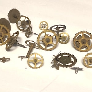 May include: A collection of vintage brass clock gears of various sizes and shapes, scattered on a white surface. The gears have intricate teeth and central holes, showcasing the mechanical components of timepieces.
