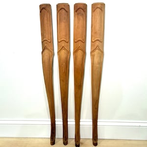 May include: Four wooden furniture legs, each with a light brown finish and a decorative carved design. The legs are tapered and stand upright against a white wall and light-colored floor.