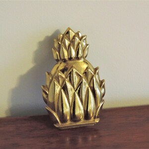 May include: A gold-colored pineapple-shaped decorative object. The pineapple has a textured surface with detailed leaf-like segments. The object is sitting on a dark wooden surface, with a neutral-colored wall in the background.