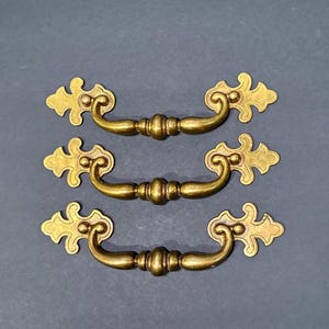 May include: Three antique brass drawer pulls with ornate floral designs. Each pull features a curved handle with a central cylindrical section and decorative end pieces. The hardware is a warm, aged gold color, suitable for furniture restoration.