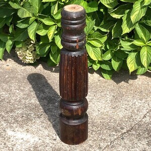 May include: A dark brown, antique wooden furniture leg. The leg has a turned design with a series of rounded and fluted sections. The wood has a rich, aged appearance, with visible grain and a slightly weathered finish. The leg is standing on a concrete surface.