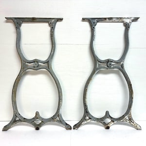 May include: Two antique, gray-painted cast iron table legs. Each leg features a decorative, open design with curved lines and ornate details. The legs are designed to support a tabletop and add a vintage aesthetic to furniture.