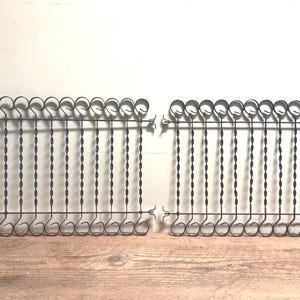 May include: Two sets of dark metal skewers, each with a decorative looped handle and a twisted shaft. The skewers are designed to hang from a horizontal bar with decorative curls at each end. The sets are displayed on a wooden surface.