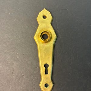 May include: A vintage brass door plate with a keyhole and mounting holes. The plate has a decorative, elongated shape with a central keyhole and a circular opening for the doorknob. The brass has a slightly aged patina.