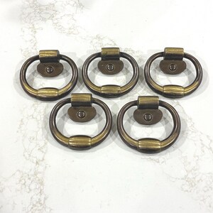 May include: Five antique brass ring pulls arranged on a white surface. Each pull features a circular ring attached to a rectangular base with a single screw hole. The brass has a slightly aged, tarnished appearance.