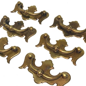 May include: A set of six brass drawer pulls, each featuring a curved, swinging handle attached to a decorative, ornate backplate with scalloped edges and a warm, metallic finish. These hardware pieces are arranged in a group on a plain surface.