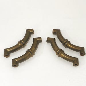 May include: Four antique brass drawer pulls. Each pull has a curved design with decorative detailing in the center and square ends. The pulls are a warm, aged brass color, suggesting vintage or retro style.