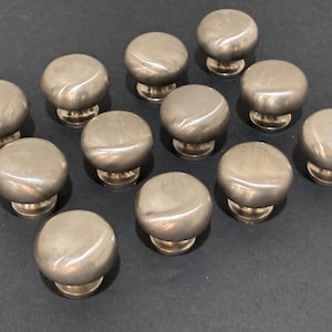 May include: A collection of twelve brushed nickel cabinet knobs. Each knob has a rounded, domed top and a short, cylindrical base. The knobs are arranged on a dark surface, showcasing their metallic sheen and simple design.
