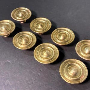 May include: A set of seven circular brass cabinet knobs arranged in a staggered pattern. Each knob features a smooth, polished gold-toned finish with a raised central dome surrounded by concentric circular ridges, creating a tiered profile.