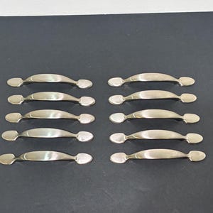 May include: Ten silver-toned cabinet handles arranged on a dark surface. Each handle has a curved bar with rounded ends, suitable for drawers or doors. The handles are made of a shiny metal, reflecting light.