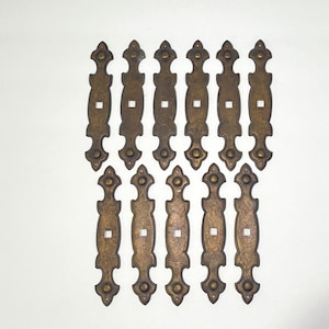 May include: A collection of antique bronze-colored cabinet pulls. Each pull has an elongated, ornate design with decorative finials and a central square opening. The hardware is arranged against a white background.
